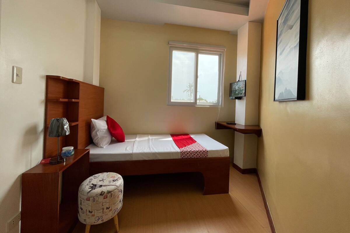 Een compacte slaapkamer met een enkel bed, een klein nachtkastje, een stoel, een raam en een aan de muur bevestigde televisie.