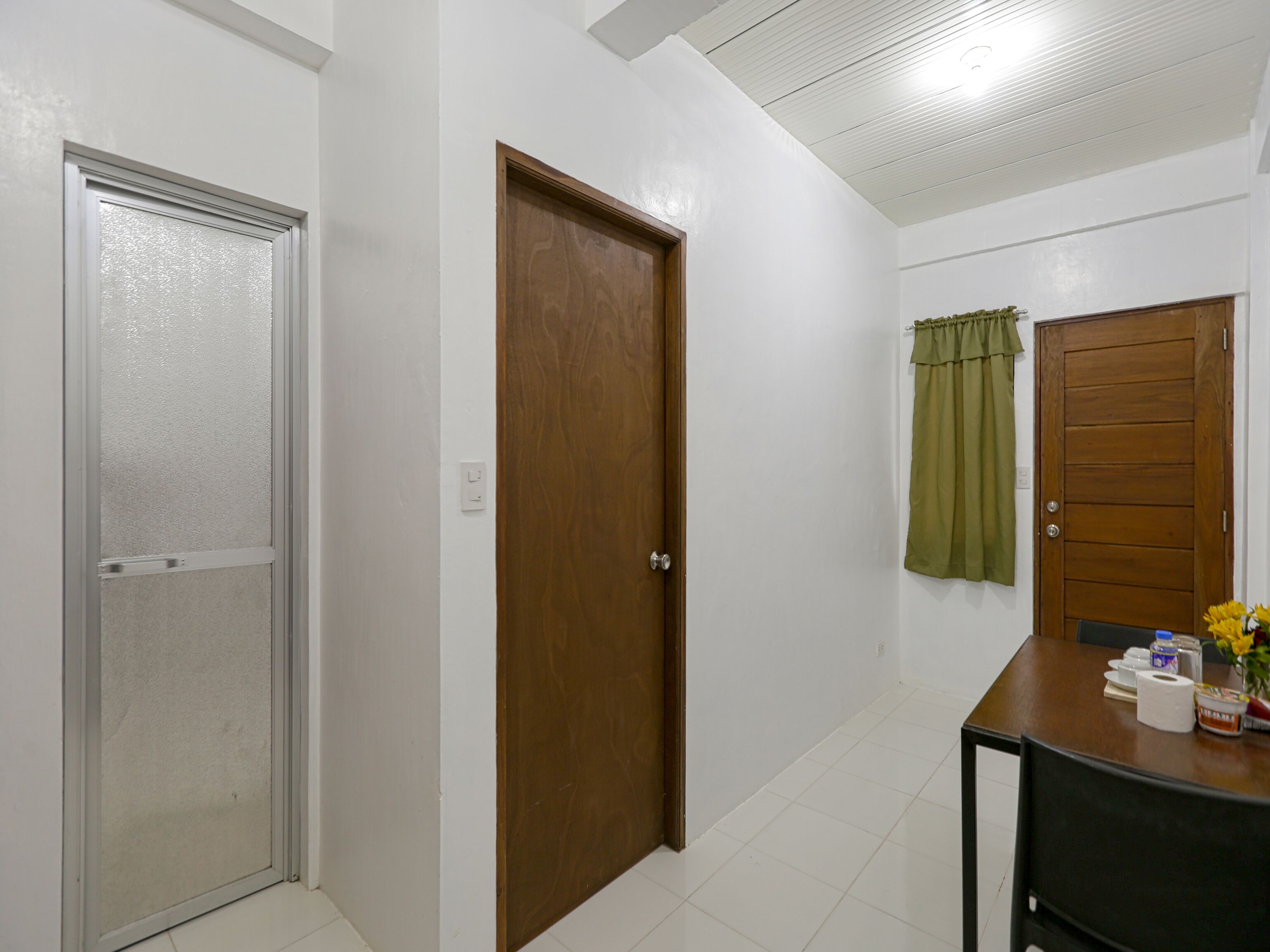 A hallway with a glass door, a wooden door, and a wooden door with a curtain. There is a table with a vase of flowers and some items on it.