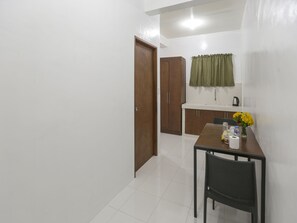 Una pequeña kitchenette con un lavamanos, armarios y una ventana con cortina verde. Hay una mesa de comedor con sillas y un florero con flores amarillas.