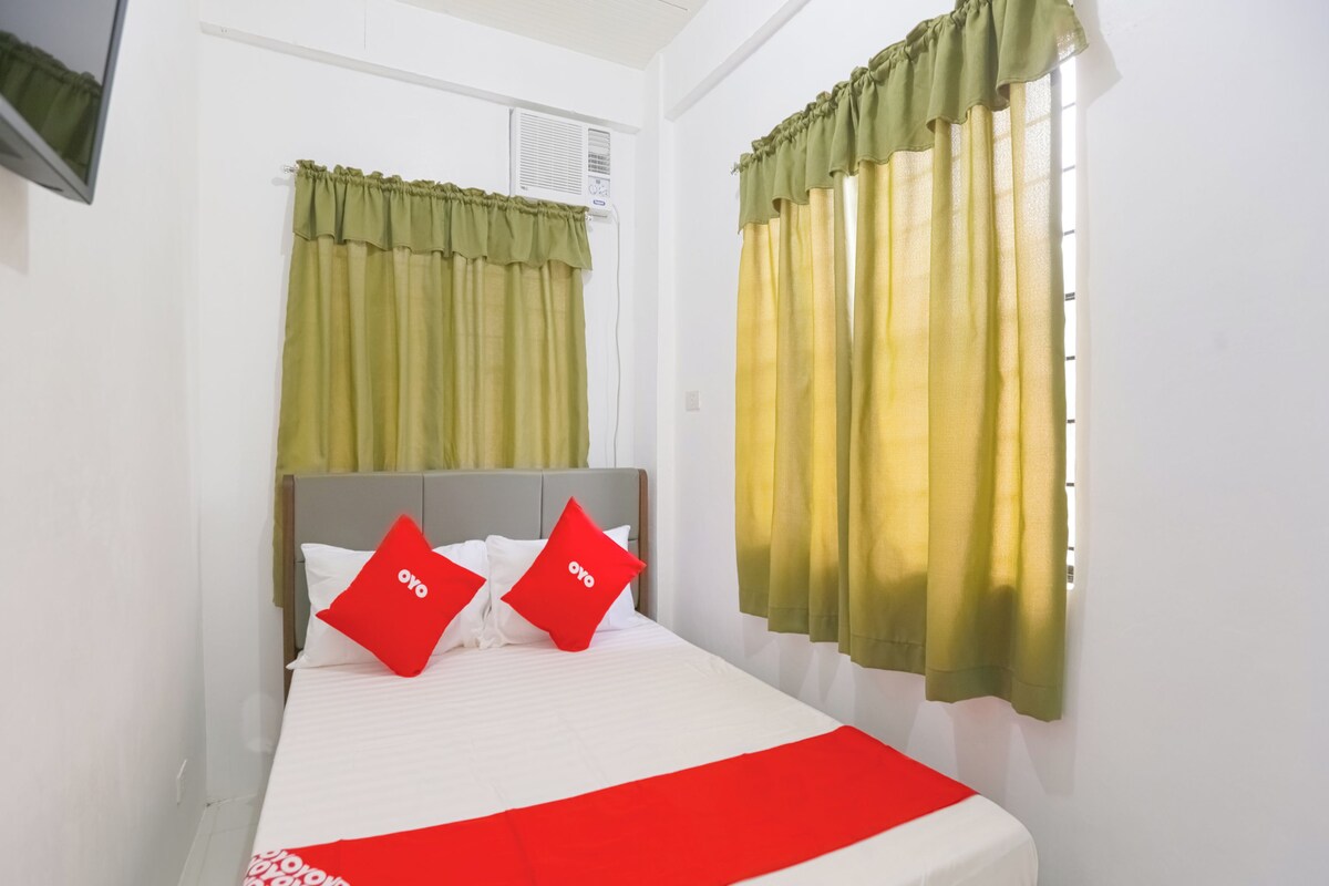 A neatly made bed with white bedding and red accents, a grey headboard, and green curtains.