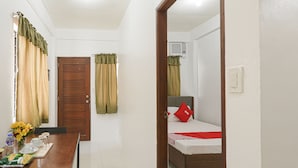 A hotel room with a wooden door, a bed with a red pillow, a small table with a vase of flowers, and a window with curtains. - OYO 736 Jade Apartelle (Cabuyao)