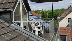 Un balcon avec une chaise blanche et un grand parasol, donnant sur un quartier résidentiel aux toits recouverts de tuiles rouges et à la végétation luxuriante.