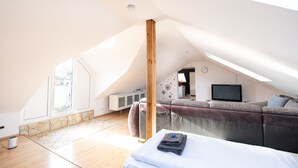 Une chambre de style loft moderne, avec un plafond en pente, une poutre en bois intégrée, une grande fenêtre, un mur d’accent en pierre, un canapé gris, un téléviseur à écran plat et un lit soigneusement fait.