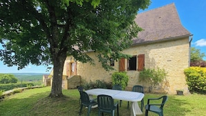Une maison en pierre avec un toit de tuiles, une table entourée de chaises et un grand arbre au premier plan.