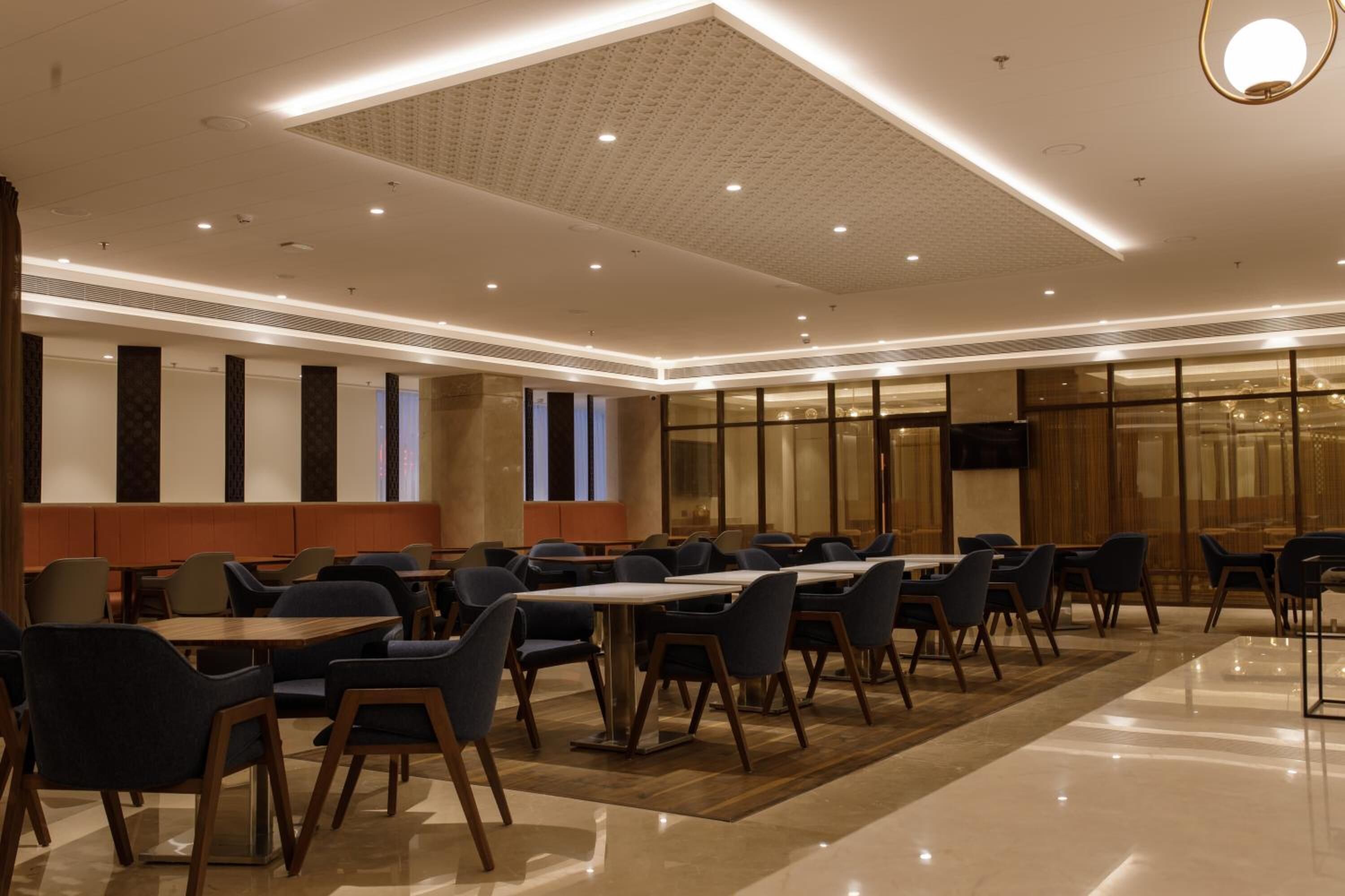 A modern hotel lobby with a long dining area, comfortable seating, and a large screen on the wall.