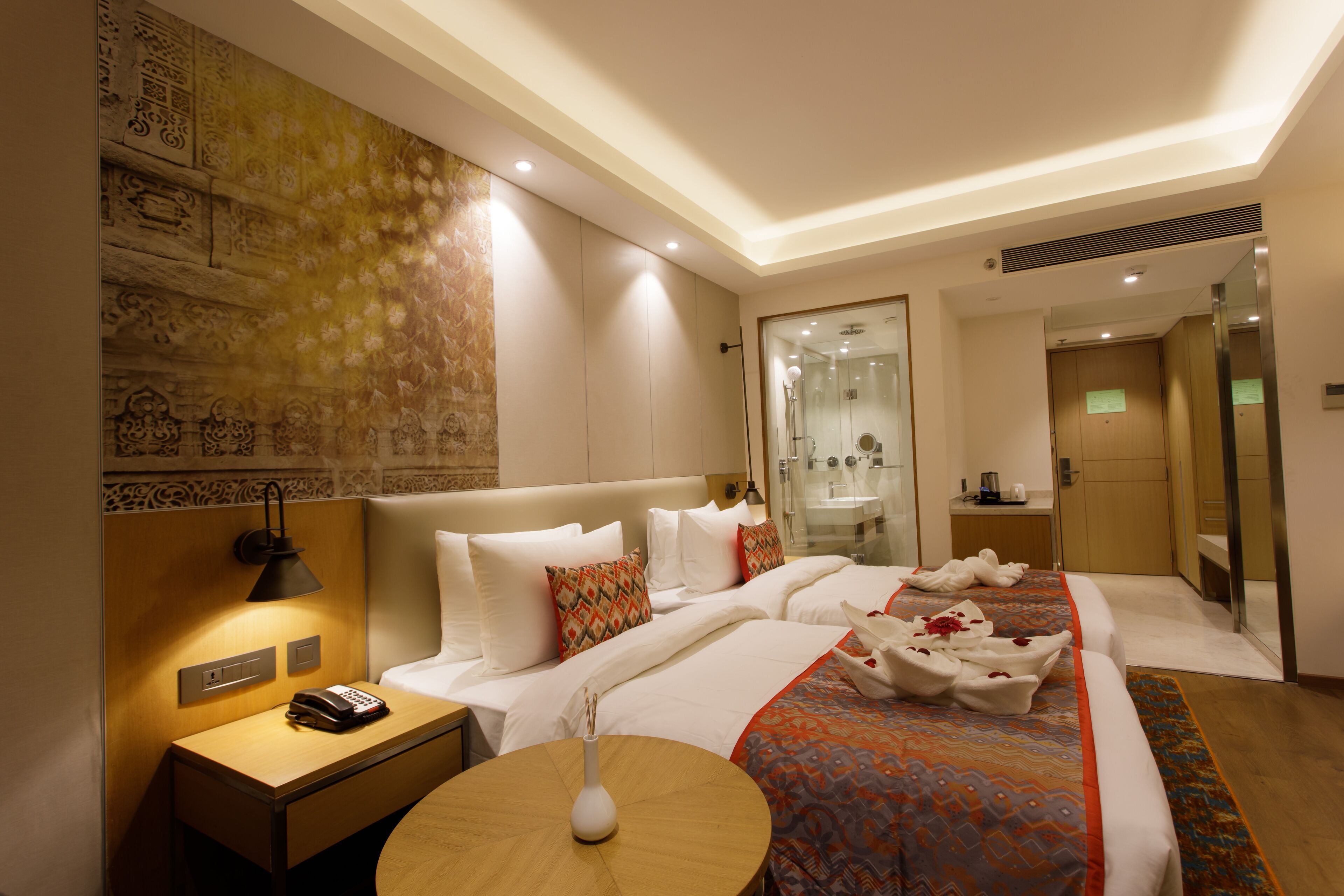 A hotel room with a large bed, a bedside table with a lamp, a small table with a vase, and a wall with a decorative pattern.