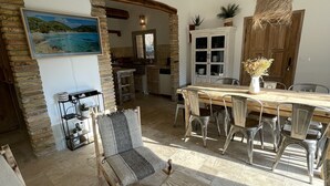 Een ruime woonkamer met een eettafel, stoelen en een bank. Achtergrond is een keuken met een houten deur en kastjes.