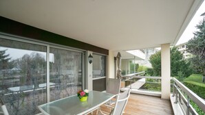 Ein moderner Balkon mit Glastisch und -stühlen, einem Blumentopf und Blick auf die umliegende Bepflanzung und Gebäude.