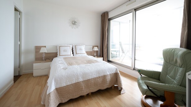 Ein Schlafzimmer mit einem großen Bett, einem Nachttisch, einem grünen Sessel und einem großen Fenster.