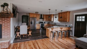 A modern kitchen with wooden cabinets, a central island with stools, a brick fireplace, and a dining area with a table and chairs. - Salmon Gathering Place sleeps 35 + perfect for reunions.retreats.events (Salmon)