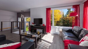 Ein modernes Wohnzimmer mit Sofa, Couchtisch und Fernseher. Ein Balkon mit Geländer bietet Blick auf Herbstbäume.