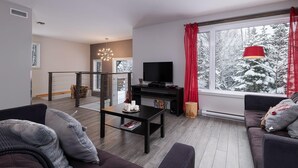 Ein modernes Wohnzimmer mit einer Couch, einem Couchtisch, einem Fernseher und einem großen Fenster mit Blick auf schneebedeckte Bäume.