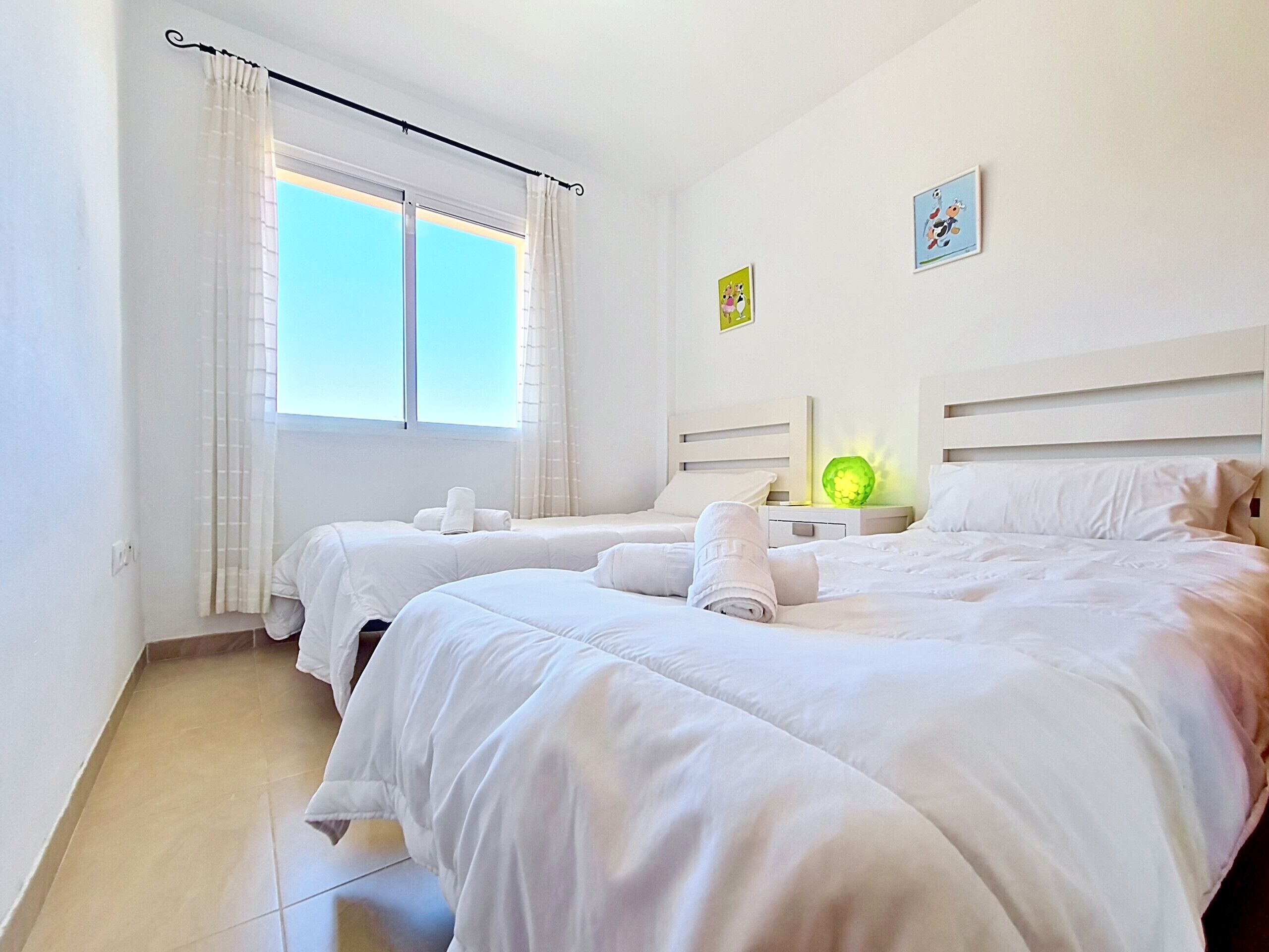 A hotel room with two beds, a window with curtains, a nightstand with a lamp, and two small wall art pieces.
