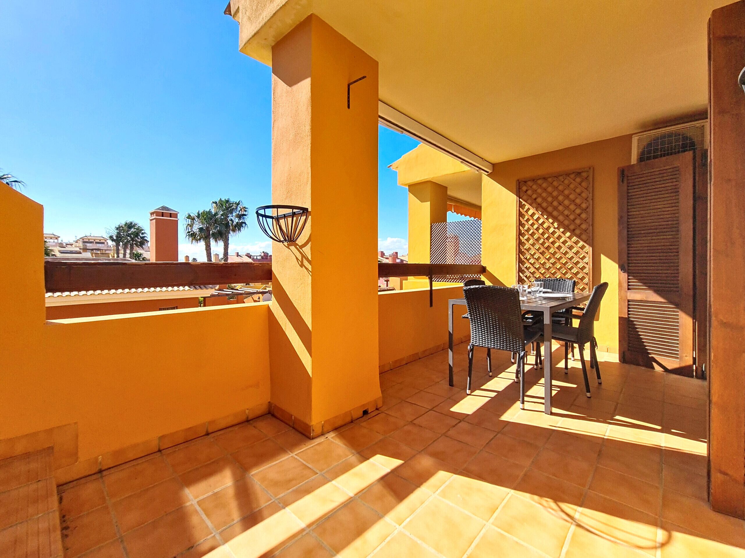 Un balcón con mesa y sillas, una canasta de baloncesto y vistas a palmeras y edificios.