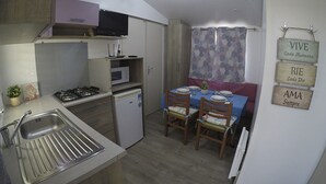 A compact kitchen with a gas stove, microwave, and sink. There is a dining area with a table set for two, chairs, and a small potted plant. A television is mounted above the stove, and there are signs on the wall with motivational phrases. - Mobile home 2 rooms (Puerto Real)