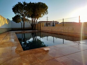 Una piscina amb paviment de rajoles, una zona tancada per una tanca i un edifici al fons.