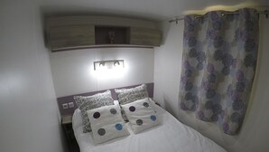 A bed with white bedding and patterned pillows, a headboard with a built-in shelf, and a curtain with a floral pattern. - Mobile home 2 rooms (Puerto Real)