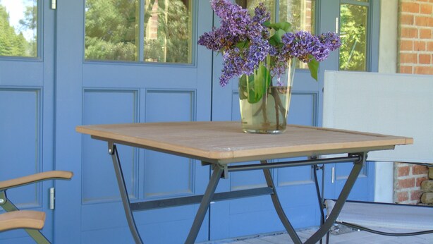 Une table sur laquelle est posé un vase rempli de fleurs violettes, devant une porte bleue ornée de fenêtres.