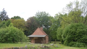 Un petit bâtiment en briques, surmonté d’un toit en tuiles, entouré d’une végétation luxuriante et d’arbres.