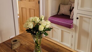 En dörr av trä, en vit bänk med en lila kudde, en liten hylla med en regnbågsfärgad dekoration och en vas med vita blommor på ett träbord.