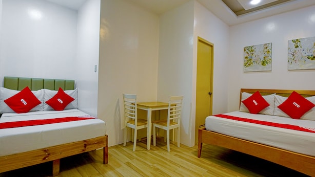 A room with two beds, each with red pillows and a white bedspread, a small white table, and a chair. There are two paintings on the wall and a door in the background. - Freemont Place (Parañaque)