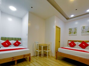 Habitación con dos camas, cada una con almohadas rojas y una colcha blanca, una mesita pequeña y una silla. Hay dos cuadros en la pared y una puerta al fondo.
