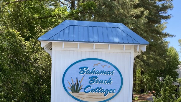 Schild für 'Bahamas Beach Cottages' mit blauem Dach und weißen Wänden, umgeben von Kies und Grünpflanzen.