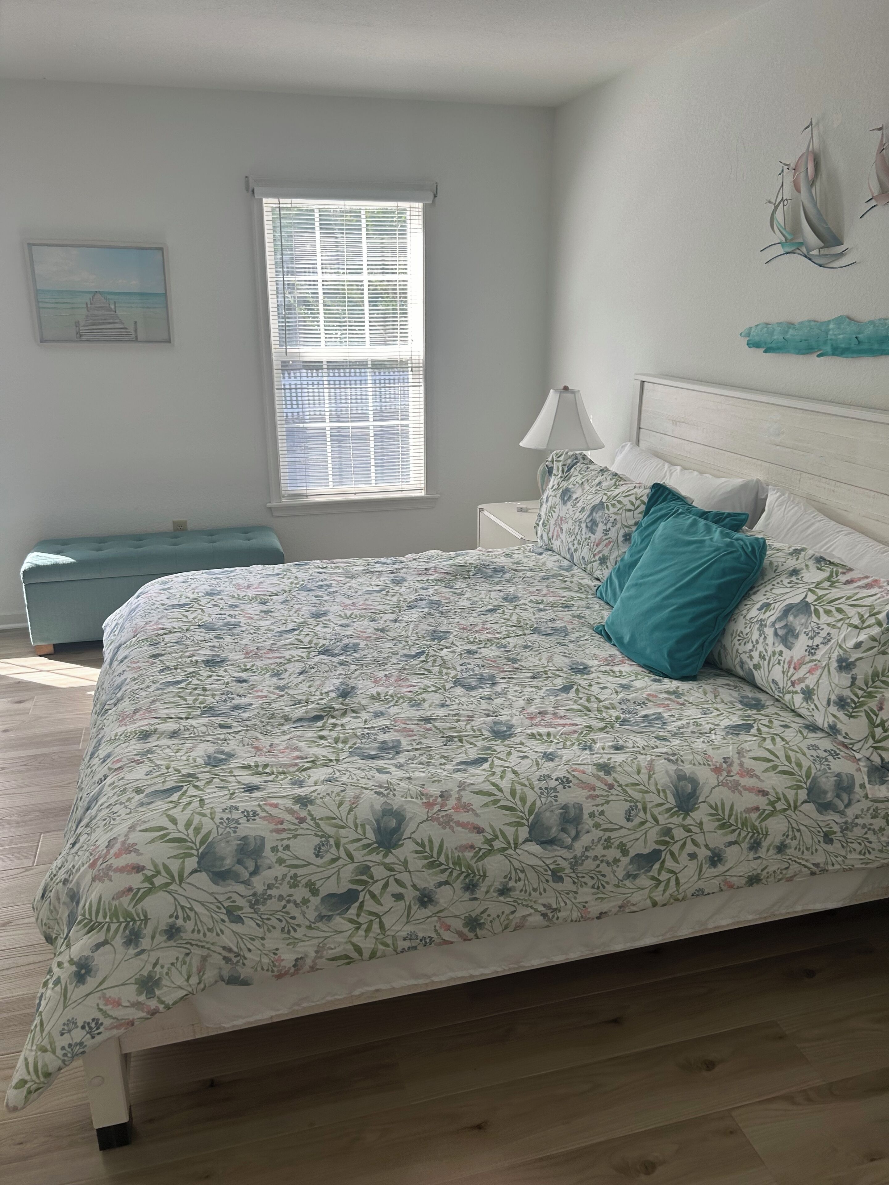 A bedroom with a bed, a nightstand, a window with blinds, a bench, and wall art.