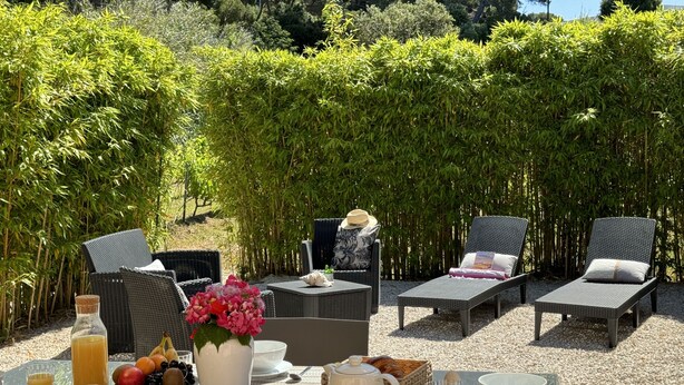 An outdoor dining area with wicker furniture, a table set with breakfast items, and a backdrop of bamboo plants. - Auguière East, house in the heart of the vineyards (Hyères)