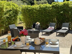 An outdoor dining area with wicker furniture, a table set with breakfast items, and a backdrop of bamboo plants.