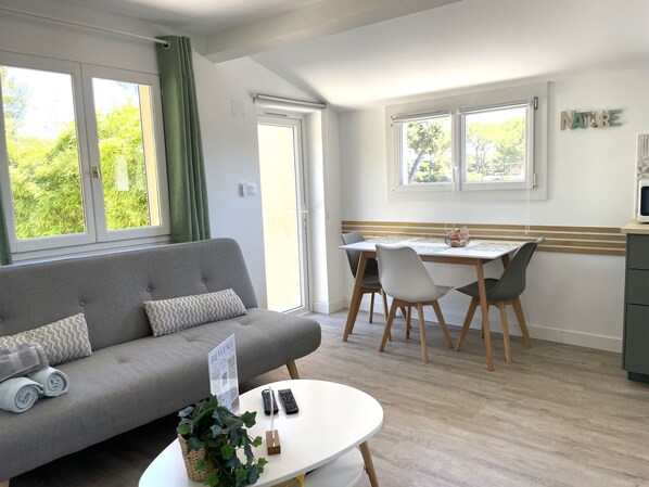 A modern living room with a grey sofa, a dining table with chairs, and a small kitchen area.