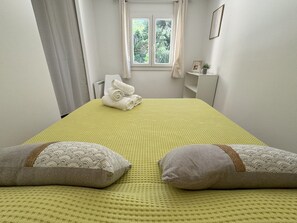 A neatly made bed with a yellow textured bedspread, two pillows with a patterned design, and a window with curtains in the background.