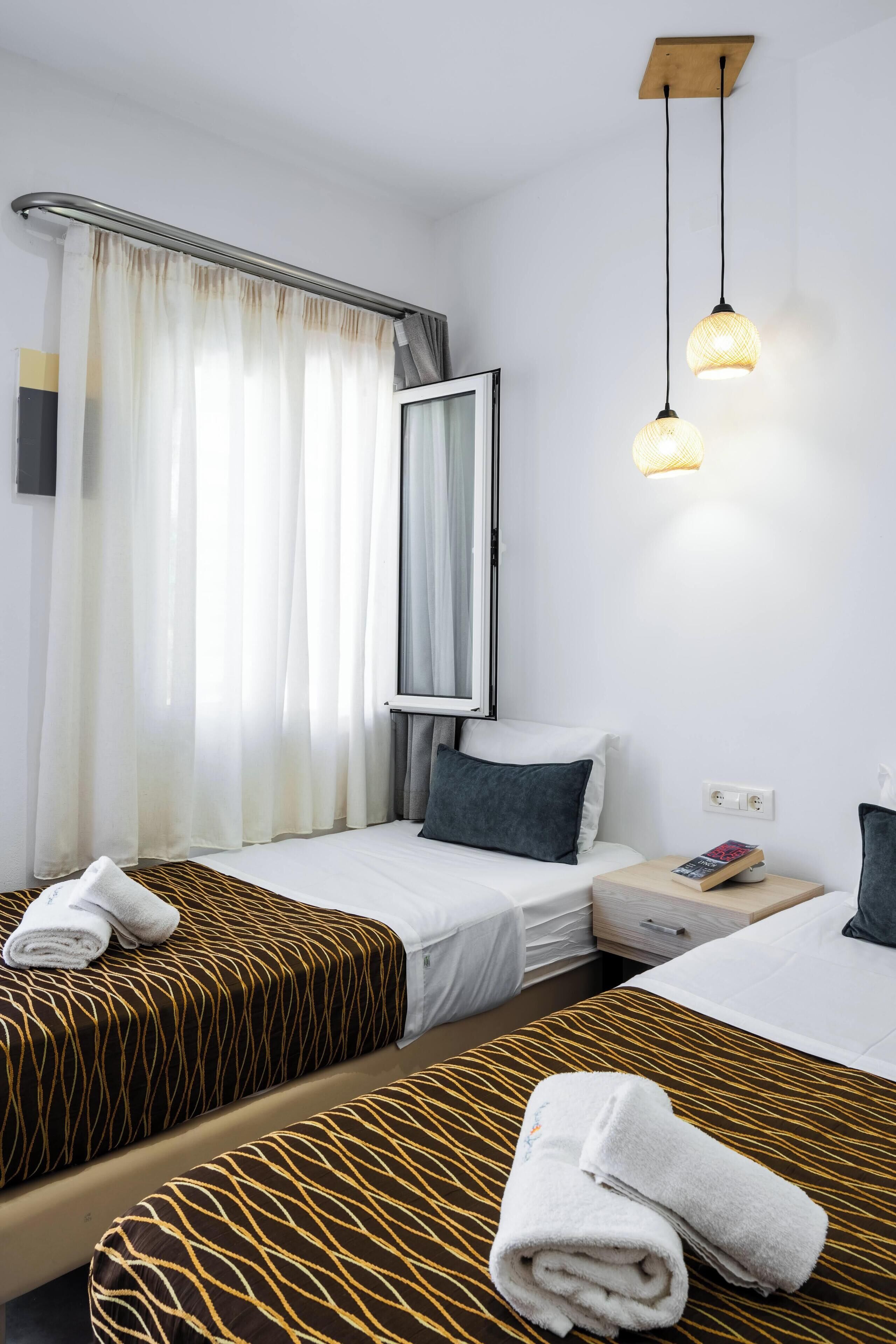 A hotel room with two beds, a bedside table, a nightstand, a mirror, and a window with curtains.