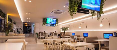 Un restaurante moderno con mobiliario blanco, plantas colgantes y varias pantallas mostrando contenido.