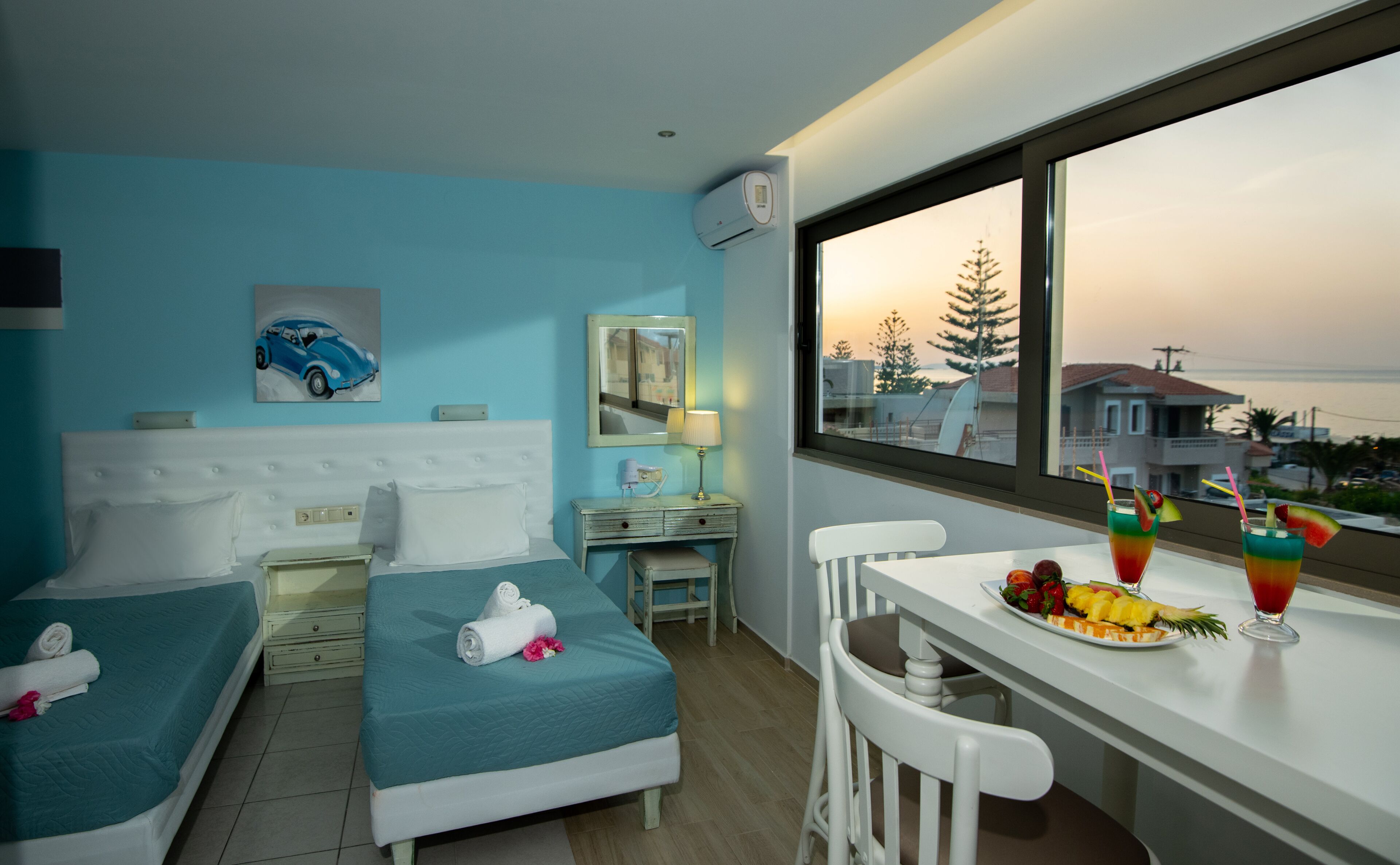 A hotel room with two beds, a desk with a chair, a window with a view of the sea, and a table with fruit and drinks.