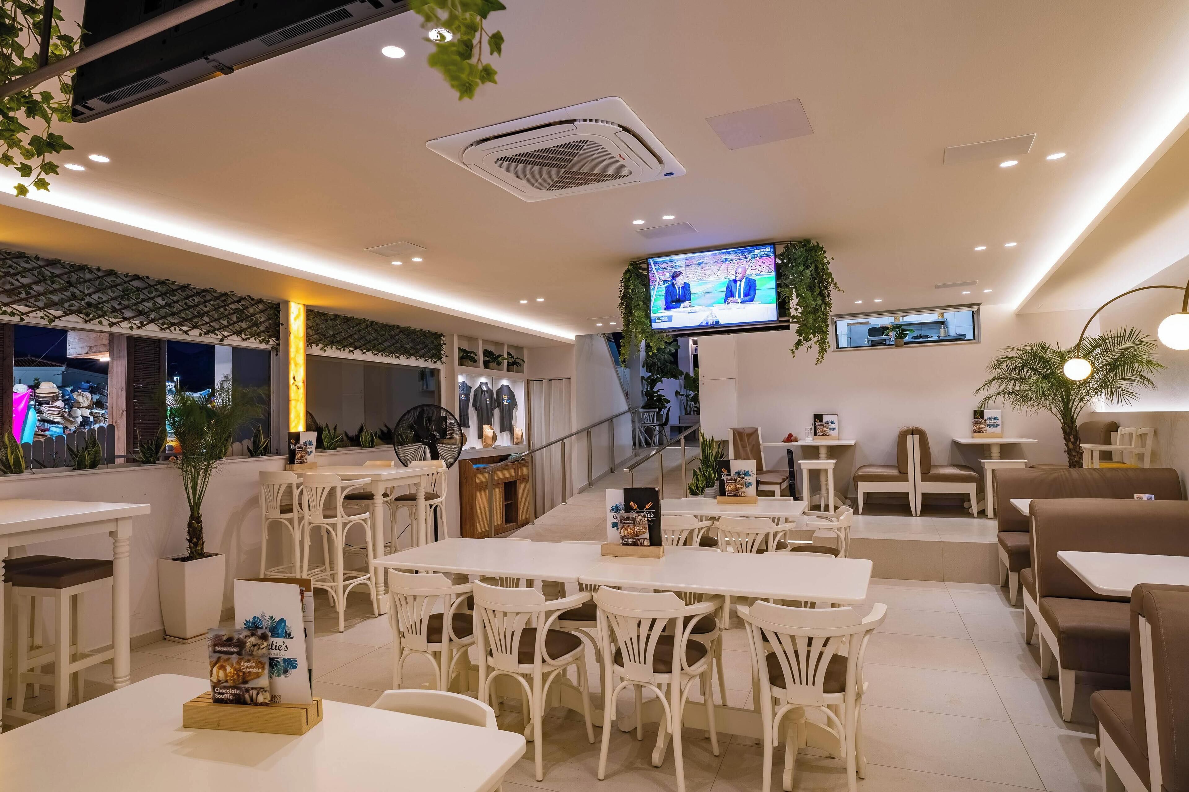 A modern restaurant with white furniture, a TV screen, and a staircase leading to an upper level.