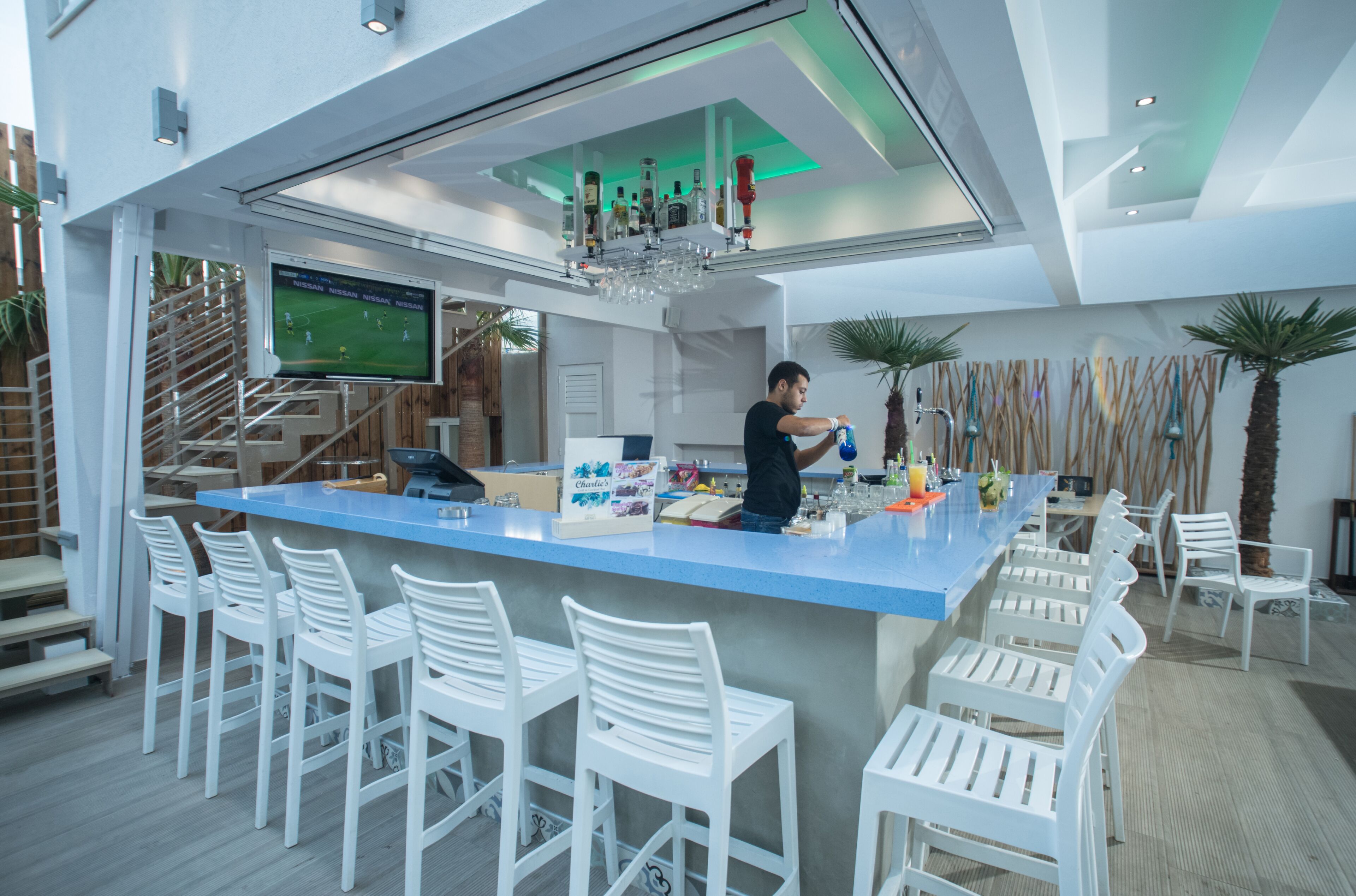 A modern bar area with a blue countertop, white bar stools, and a television screen displaying a sports game.