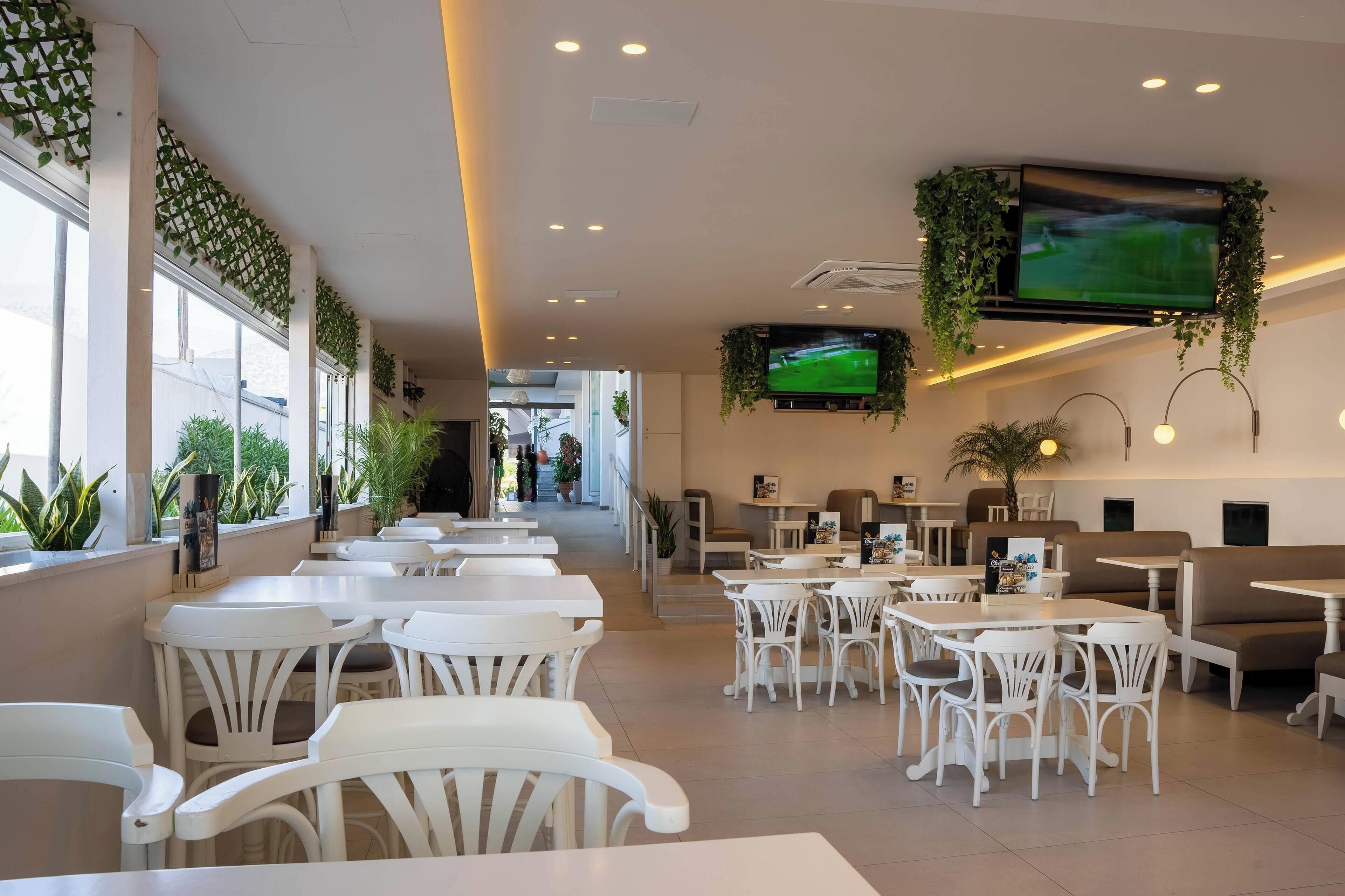 Un espacio interior amplio con mesas y sillas blancas, un televisor grande montado en la pared y plantas colgando del techo.