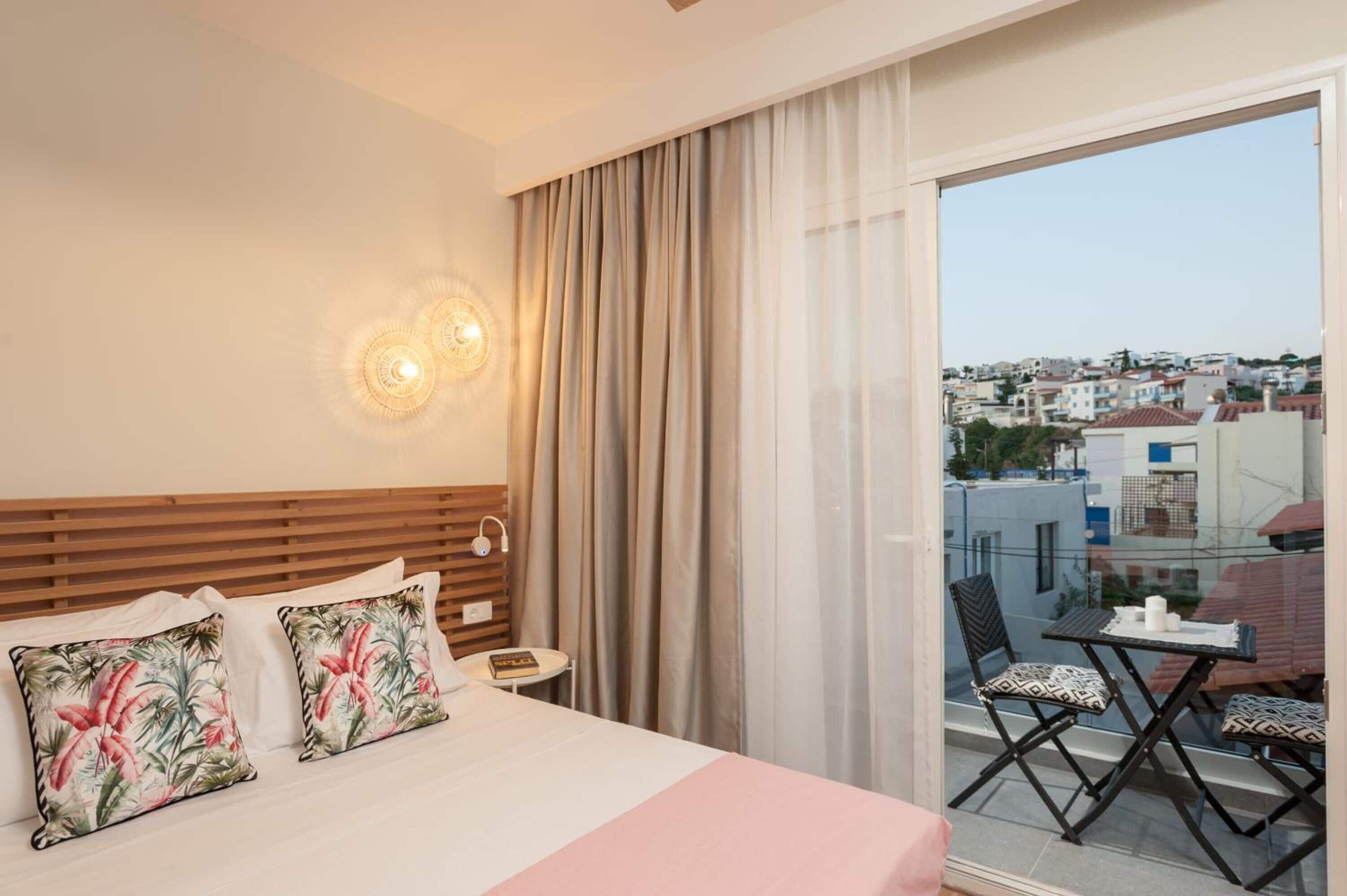 A hotel room with a bed, a small table with chairs, a nightstand, and a view of the city through a balcony.
