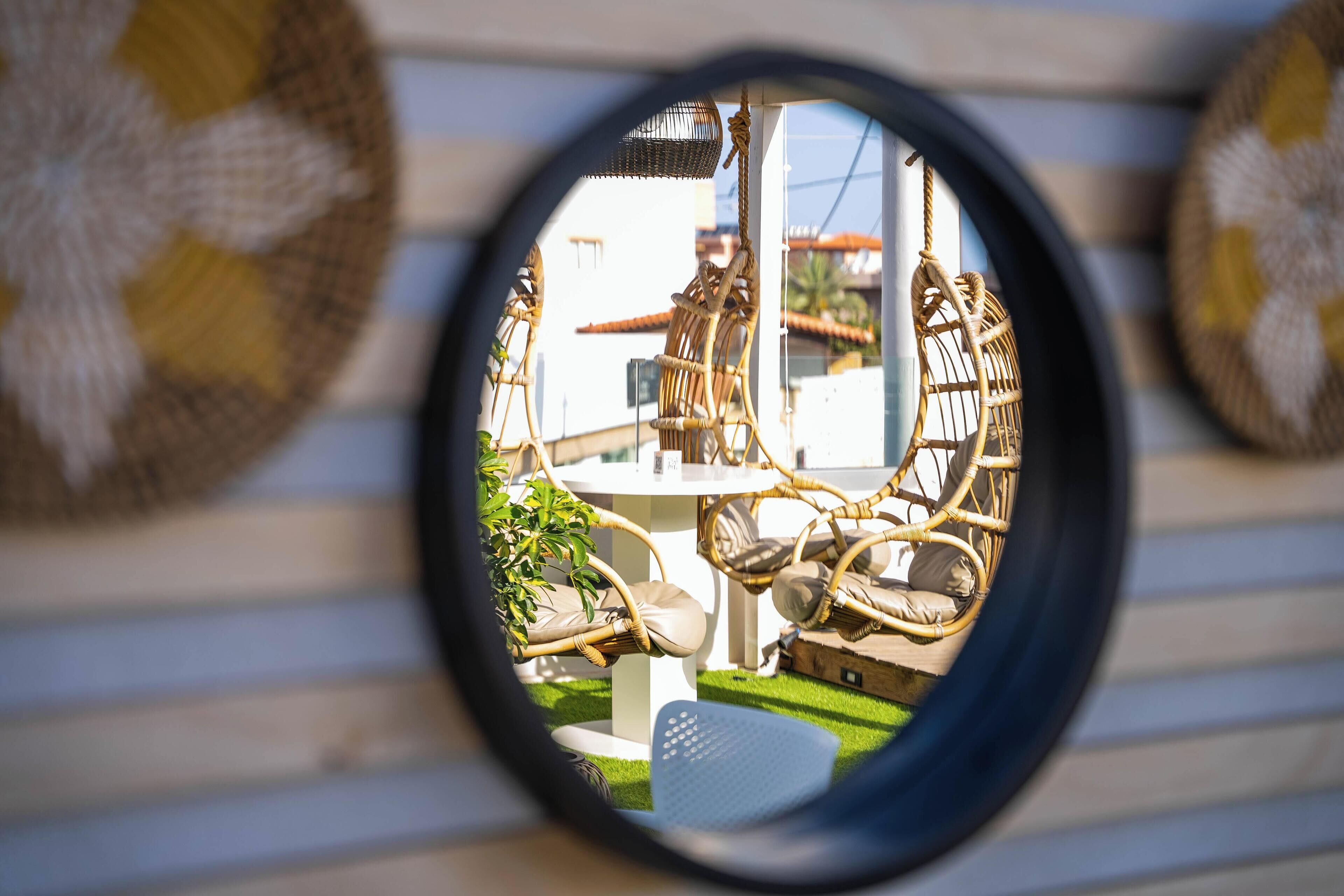 Un espejo redondo que refleja muebles de mimbre y una mesa blanca, evocando un espacio de estar al aire libre.