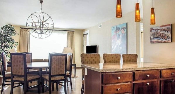 Habitación de hotel con zona de comedor, una isla grande con taburetes de bar, una araña, un televisor de pantalla plana y un cartel en la pared.