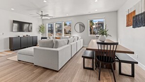 Een moderne woonkamer met een grijze hoekbank, een houten eettafel met stoelen, een plafondventilator en een groot raam met uitzicht.