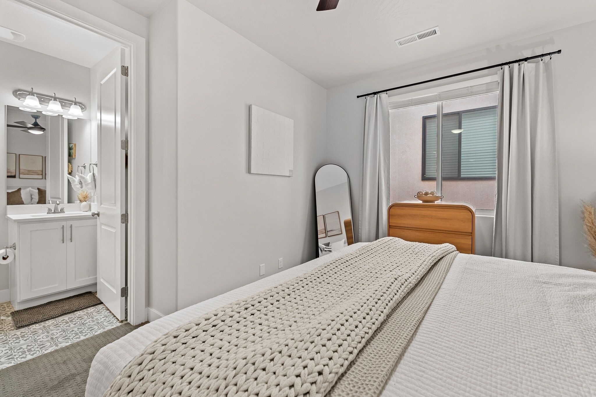 A bedroom with a bed, a dresser, a mirror, a window with curtains, and a bathroom with a vanity and mirror.
