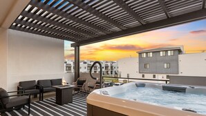 Een terras op het dak met een jacuzzi, een zitgedeelte en uitzicht op de ondergaande zon boven een woongebied.