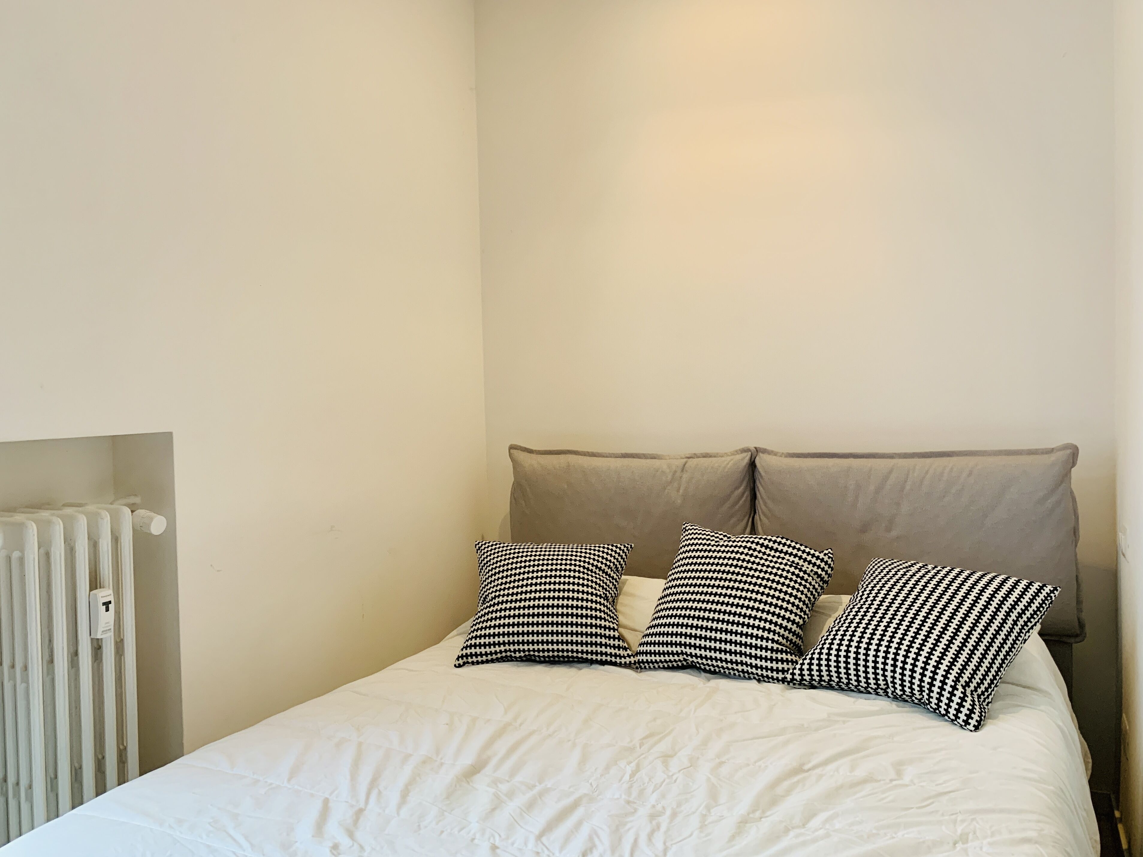 A bed with a white duvet and three black and white patterned pillows, a beige headboard, and a white radiator on the left.