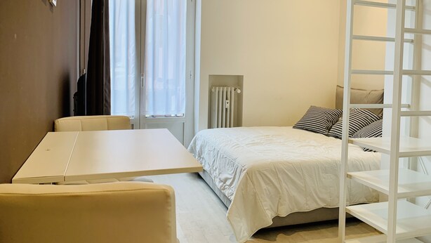 A compact hotel room with a bed, a small table, a chair, a radiator, and a window with curtains.