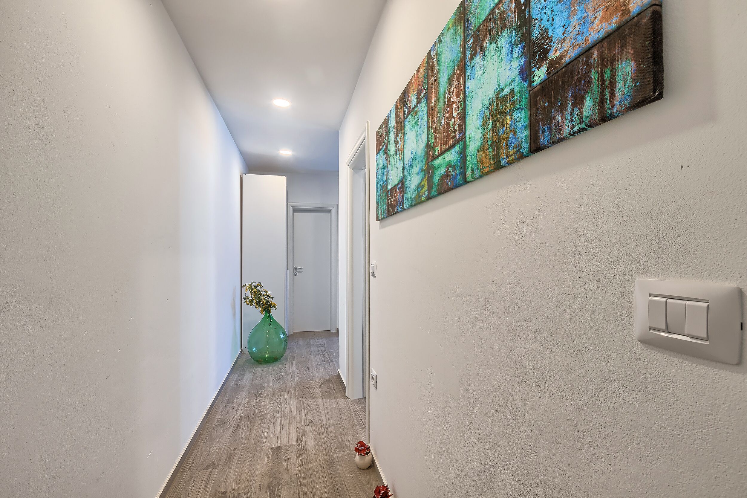 A hallway with a wooden floor, a green vase, and a painting on the wall.