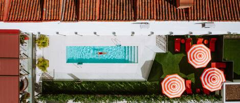 An aerial view of a rooftop pool area with a swimming pool, red umbrellas, and surrounding greenery.