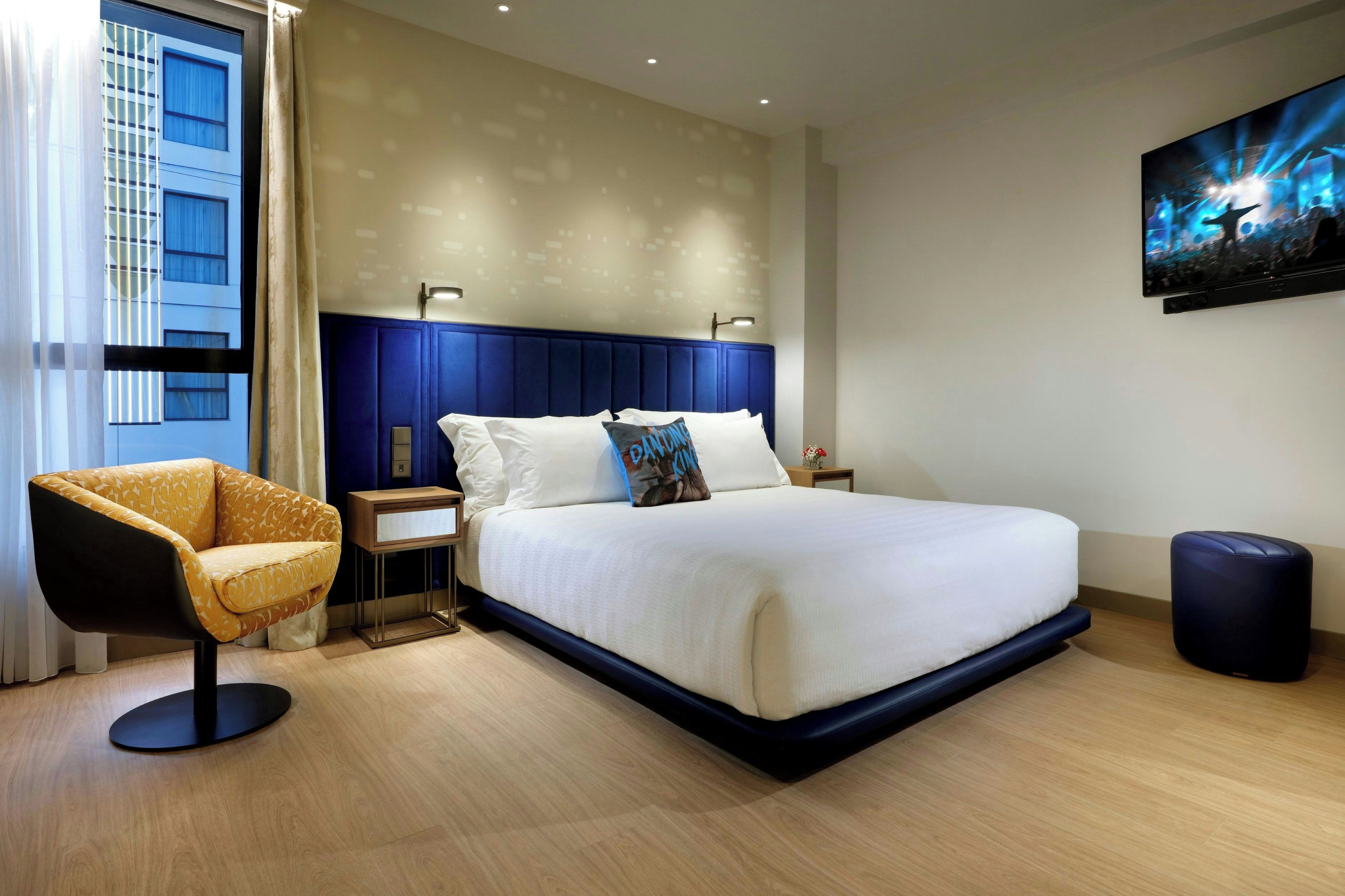 Una habitación de hotel moderna con una cama grande, una mesita auxiliar, una silla y un cuadro en la pared.