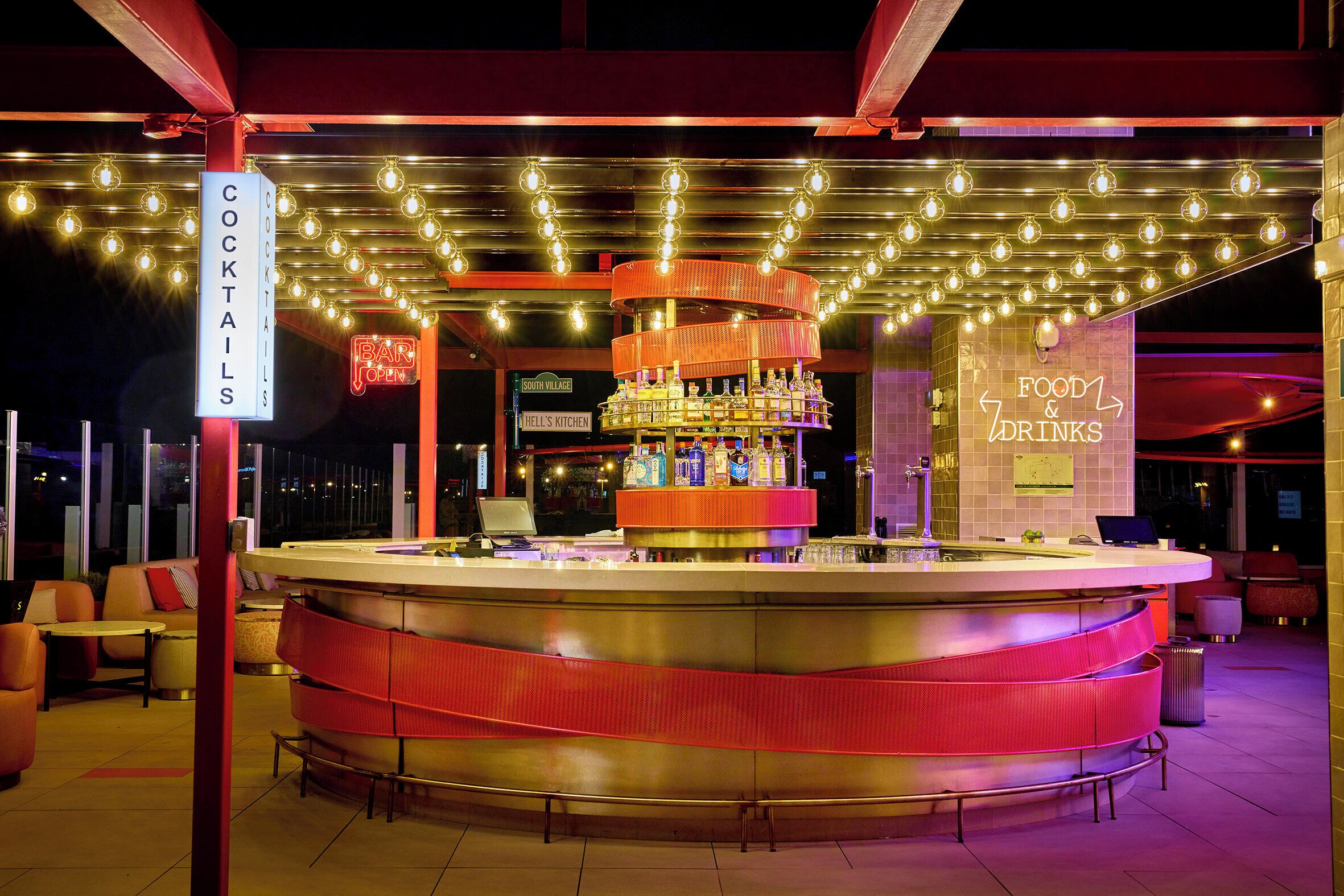 Un espace bar doté d’un comptoir circulaire, de nombreuses bouteilles et d’un panneau indiquant les cocktails proposés.